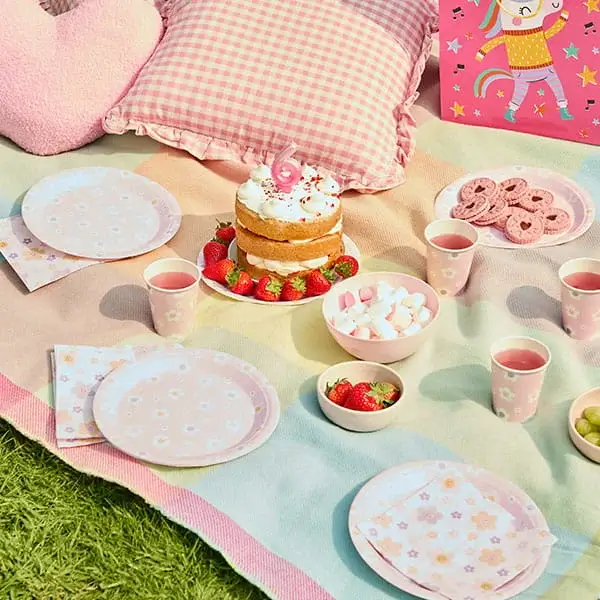 Pastel picnic party setup with a birthday cake, strawberries, snacks, pink tableware, cushions, and a picnic blanket on the grass.